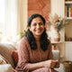 A smiling South Asian parent posing for a professional headshot in a bright, modern room with warm terracotta and cream accen