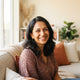 A smiling South Asian woman in a bright, modern living room with warm cream and terracotta accents.